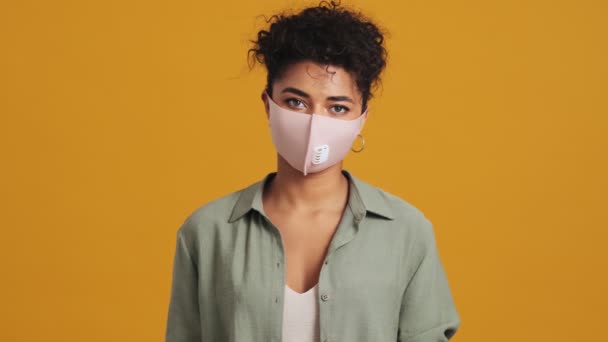 幸せなアメリカ人女性のクローズアップビューは スタジオで黄色の壁に隔離された親指アップジェスチャーを行う顔の医療保護マスクを身に着けています — ストック動画