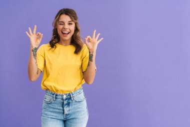 Portrait of a beautiful excited young casual woman standing isolated over blue background, showing ok and winking clipart