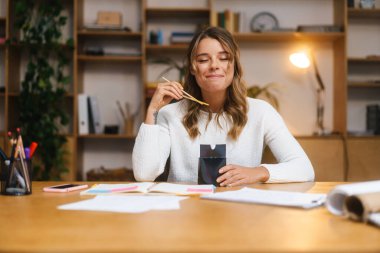 Gülümseyen genç girişimci kadın ofis masasında oturmuş erişte yerken öğle yemeği yiyor.