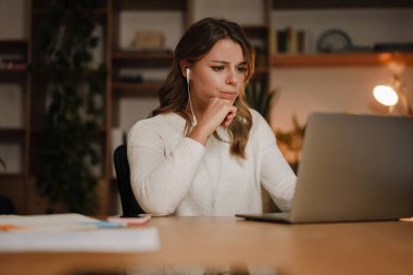 Kulaklıklı ciddi bir iş kadını dizüstü bilgisayarda webinar izleyip not alıyor, bilgisayar dersleri öğreniyor, arama yapıyor, çevrimiçi konferansa katılıyor, çevirmenlik eğitimi veriyor.