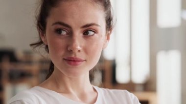 Tişörtlü gülümseyen kadının yan tarafa baktığı ve ofiste dururken kameraya baktığı yakın çekim görüntüsü.