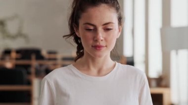 Tişörtlü gülümseyen kadın ofiste dikilirken kameraya gözlerini açıyor.