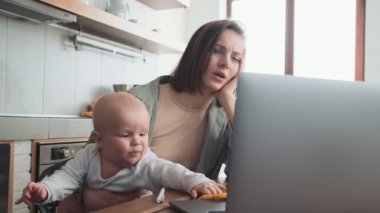 Yorgun annenin laptopta bir şey ararken ve mutfakta ağlayan bir bebeği tutarken yakın plan görüntüsü.