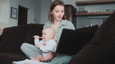 Gülümseyen bir anne dizüstü bilgisayara bakar ve kanepede otururken bebeğini emer.
