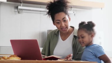 Gülümseyen Afrikalı bir anne kâğıt üzerinde kalemle bir şey gösteriyor ve mutfaktaki küçük kızına bir not defteri gösteriyor.