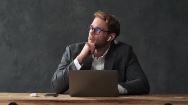 Kablosuz kulaklıklar ve ofis kıyafetiyle üzgün bir adam laptopa bakıyor ve gri stüdyodaki ahşap masada otururken kafasını tutuyor.