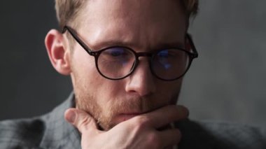 Ofis kıyafetli, gözlüklü, düşünceli bir adamın yakın plan bir resmi. Gri bir ofiste, ileri ve yana bakıyor.