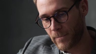 Ofis takımlı, gözlüklü ve gri bir ofiste dudaklarını ısıran dalgın bir adamın yakın plan görüntüsü.