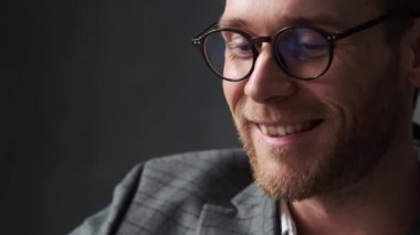 Ofis takımlı ve gözlüklü, gri bir ofiste ileriye bakan gülümseyen bir adamın yakın plan fotoğrafı.