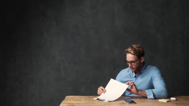 一个戴眼镜 身穿蓝色衬衫的沉思的男人 在灰色的办公室里坐在桌子旁边 一边翻着纸一边看一边看 — 图库视频影像