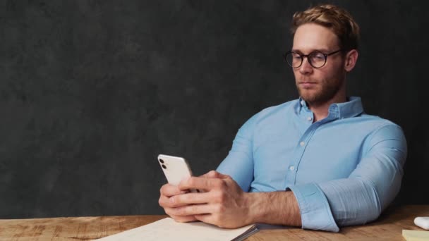 灰色のオフィスの木製のテーブルに座っている間 笑顔で携帯電話に何かを書く青いシャツに集中した男 — ストック動画
