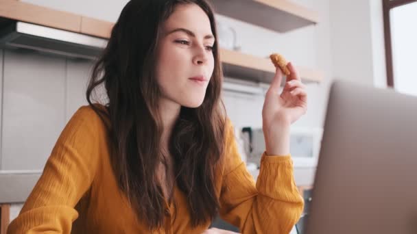 笑顔の女性がクッキーを食べ キッチンでラップトップを見て — ストック動画