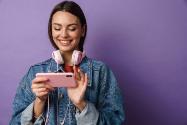 Kulaklık takan mutlu genç bir kadın, cep telefonunu mor arka planda tutuyor, ekrana bakıyor.