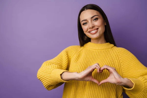 Happy young woman in sweater showing heart symbol with hands over violet wall background