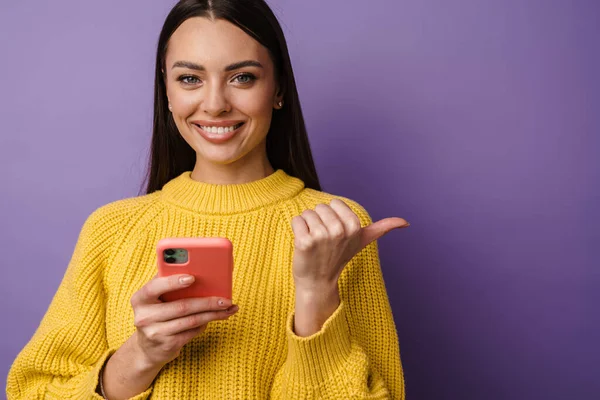 Счастливая Молодая Женщина Свитере Держит Мобильный Телефон Фиолетовом Фоне Указывая — стоковое фото