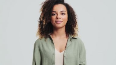 Afro-kıvırcık saçlı Afro-Amerikalı olumlu bir kadın iki eliyle baş parmak hareketi yapıyor. Stüdyoda beyaz bir duvarın üzerinde tek başına duruyor.