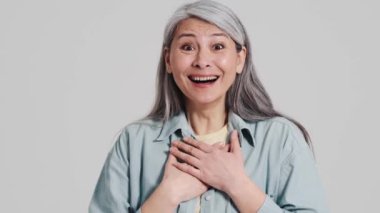 Memnun bir yaşlı kadın kameraya bakıyor. Stüdyoda gri bir duvarın üzerinde duruyor.