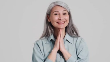 Memnun bir yaşlı kadın kameraya bakıyor. Stüdyoda gri bir duvarın üzerinde duruyor.