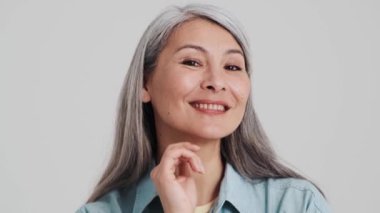 Çekici yaşlı bir kadın kameraya poz veriyor. Stüdyodaki gri bir duvarın üzerinde tek başına duruyor.