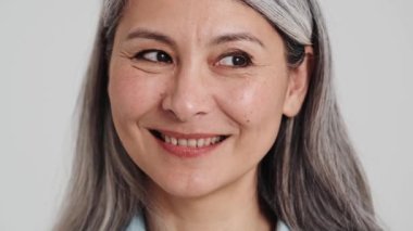 Mutlu yaşlı bir kadının yakın çekim görüntüsü stüdyodaki gri bir duvarın üzerinde yalnız başına kameraya poz veriyor.