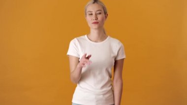 Turuncu bir stüdyoda dikilirken sessizlik jesti yapan tişörtlü, pozitif sarışın Asyalı bir kadın.