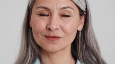 Gülümseyen yaşlı bir kadının yakın çekim görüntüsü stüdyodaki gri bir duvarın üzerinde izole bir şekilde duran kameraya sessiz bir jest gösteriyor.