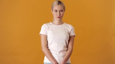 Alnını buruşturan tişörtlü, üzgün, Asyalı bir kadın turuncu bir stüdyoda dikilirken çapraz kollarıyla hareket göstermiyor.