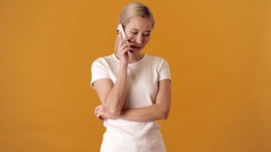 Tişörtlü, alarmlı sarışın Asyalı bir kadın turuncu bir stüdyoda dururken telefonla birini arıyor.