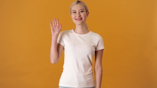 オレンジ色のスタジオに立っている間にカメラを振ってTシャツに肯定的なブロンドアジアの女性 — ストック動画