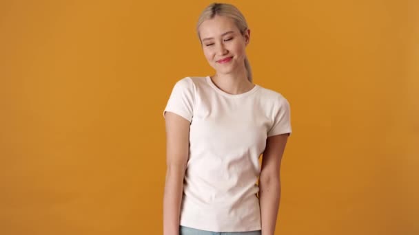 一位穿着T恤衫的快乐的金发亚洲女人 站在橙色的工作室里尴尬地低头看着 — 图库视频影像