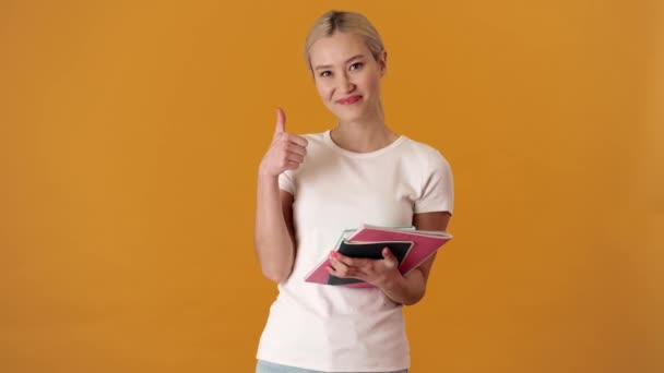 オレンジ色のスタジオで親指のジェスチャーを示す手にノートのスタックとTシャツの笑みを浮かべてアジアの女性 — ストック動画
