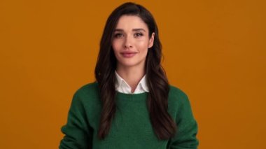 Yeşil kazaklı ve tişörtlü gülümseyen genç bir kadın turuncu bir stüdyoda dururken selam veriyor.
