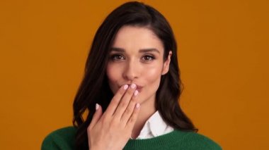 Yeşil kazaklı ve tişörtlü gülümseyen kadının turuncu bir stüdyoda canlı bir öpücük gönderişi.