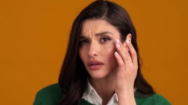 Yeşil kazaklı ve gömlekli mutsuz bir kadın aynaya bakar gibi kameraya bakıyor ve turuncu bir stüdyoda görüntüsünü düzeltiyor.