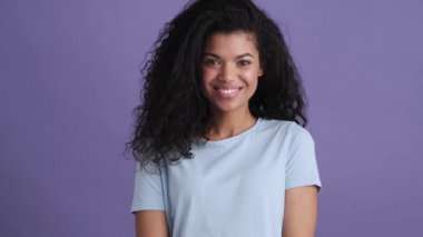 Mor bir stüdyoda tişörtlü gülümseyen kıvırcık Afrikalı bir kadın.