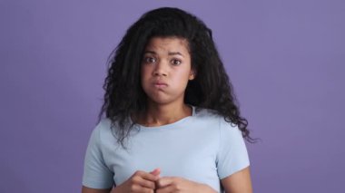 Üzgün, kıvırcık saçlı, tişörtlü bir Afrikalı kadın mor bir stüdyoda dikilirken bir şey düşünüyor.