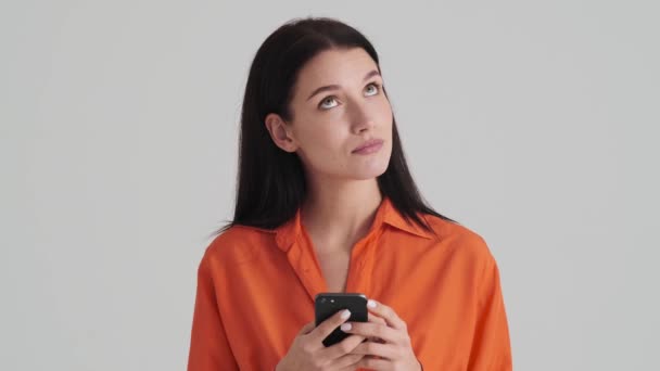 思考の女性は彼女のスマートフォンを使用しているスタジオで灰色の壁に隔離された立っている — ストック動画