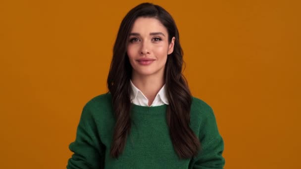 一位穿着绿色毛衣和衬衫 面带微笑的年轻女子站在橙色工作室里打招呼 — 图库视频影像