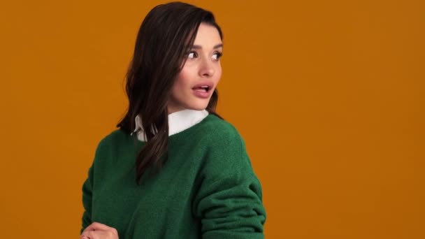 緑のセーターとシャツに身を包んだ幸せそうな女性の側面図とオレンジのスタジオに立っている間の沈黙のジェスチャー — ストック動画