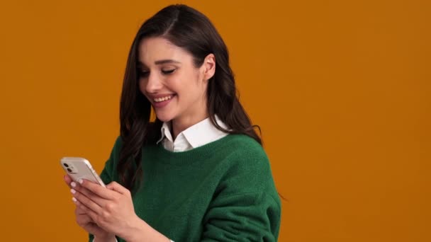 オレンジ色のスタジオに立っている間 カメラで積極的に電話を見て うなずいて緑のセーターやシャツで幸せな女性の側面図 — ストック動画