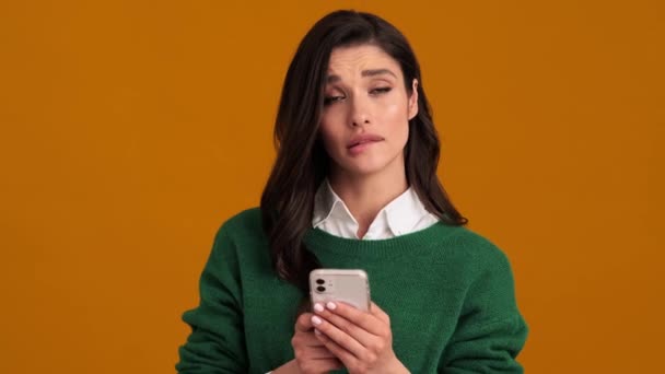 オレンジ色のスタジオに立っている間に彼女の手に電話を保持し 答えについて考えている緑のセーターとシャツの悲しい女性 — ストック動画