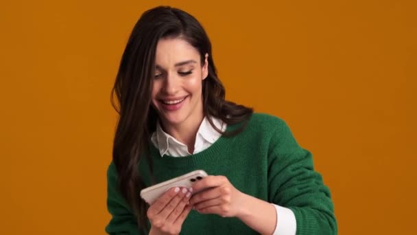 一位身穿绿色毛衣和衬衫的积极女性 一边在手机上玩游戏 一边站在橙色工作室里为自己的失利而烦恼 — 图库视频影像