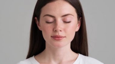 Güzel bir kadının yakın plan görüntüsü gözlerini açıyor ve stüdyonun gri arka planında tek başına duran kameraya bakıyor.