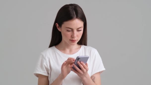 一个穿着基本T恤的快乐女人一边用她的智能手机一边指着它 一边站在工作室里灰色的背景下 — 图库视频影像