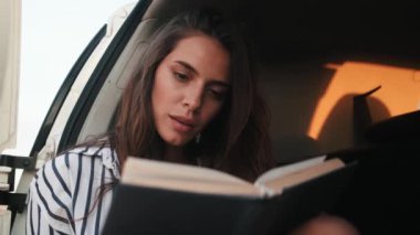 Güzel bir kadının yakın plan görüntüsü arabanın bagajında oturmuş kitap okuyor.