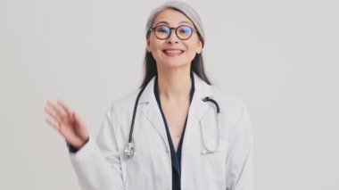 Gülümseyen kıdemli bir doktor elini kameraya doğru sallıyor. Stüdyodaki gri duvarın üzerinde tek başına duruyor.