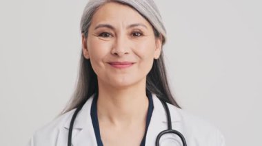 Güzel bir kıdemli kadın doktor stüdyodaki gri duvarın üzerinde dikiliyor.
