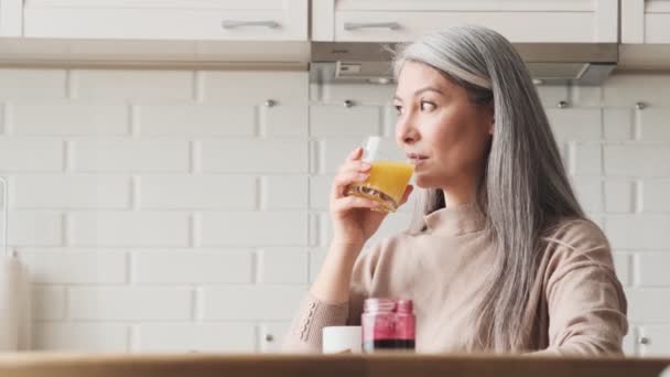 Lycklig Äldre Kvinna Dricker Ett Glas Apelsinjuice Sittandes Köket Hemma — Stockvideo