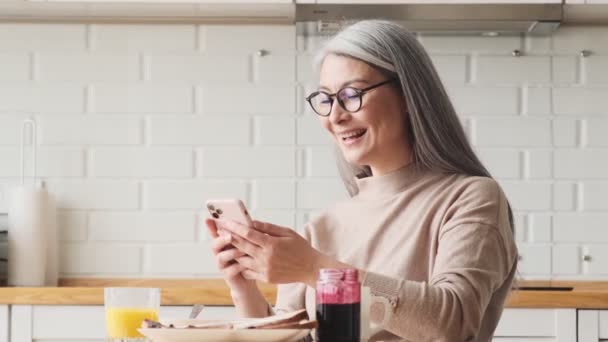 Skrattande Äldre Kvinna Använder Sin Smartphone Sittandes Köket Hemma — Stockvideo