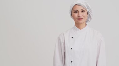 Gülümseyen bir aşçı kadın, stüdyodaki gri duvarın üzerinde izole edilmiş bir şeyi sunarken kendine güvenen bir poz veriyor.